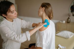A physiotherapist applies blue kinesiology tape to a patient's shoulder for muscle support.