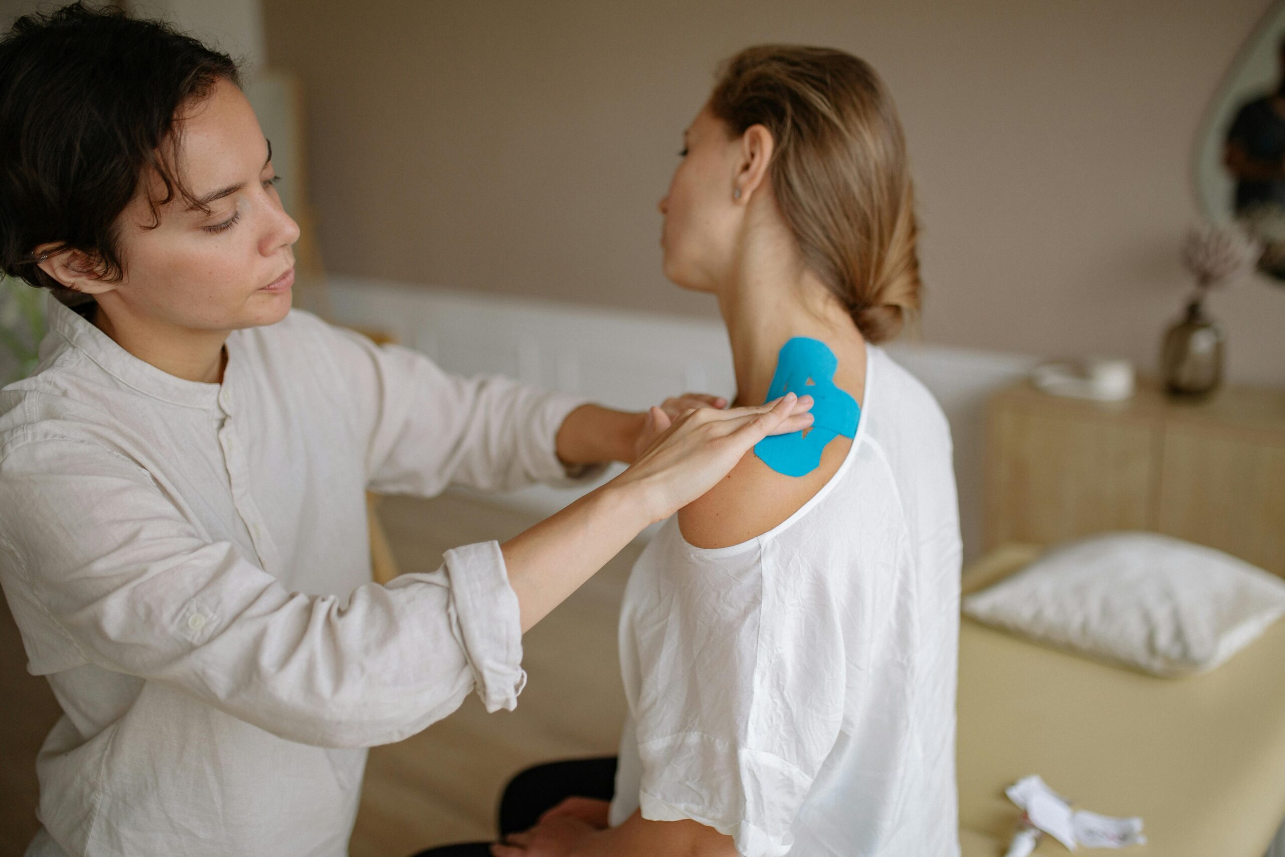A physiotherapist applies blue kinesiology tape to a patient's shoulder for muscle support.